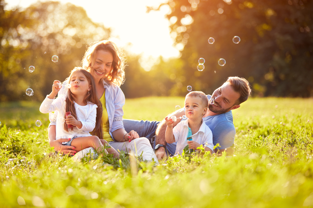 A family including a mother, father, daughter, and son enjoy blowing bubbles in a field during the summertime.